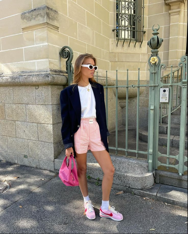 Mulher jovem loira usando blazer, camiseta básica branca, short listrado no estilo samba canção, bolsa rosa, meia branca aparente e tênis Nike Cortez retrô