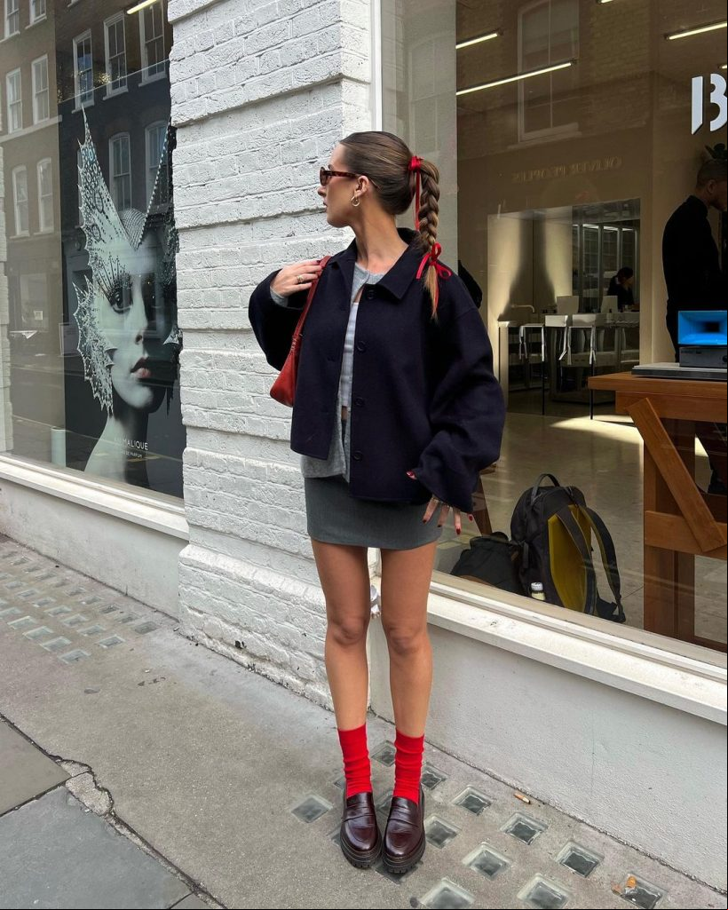 Kezia Rose Cook com laço de fita no cabelo, óculos de sol, casaco azul-marinho, cardigan cinza, regata cinza, minissaia cinza, meia vermelha e mocassim vinho